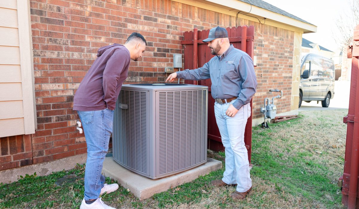 Boost Air technician inspecting Ruud outdoor AC condenser with homeowner by brick house